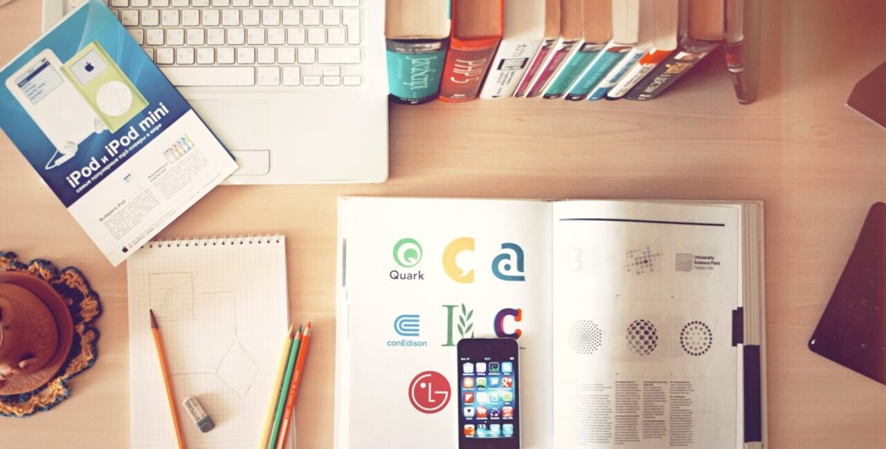 Ux web designer workplace is books, pencils, papers, a mobile phone and a white laptop on a wood desk