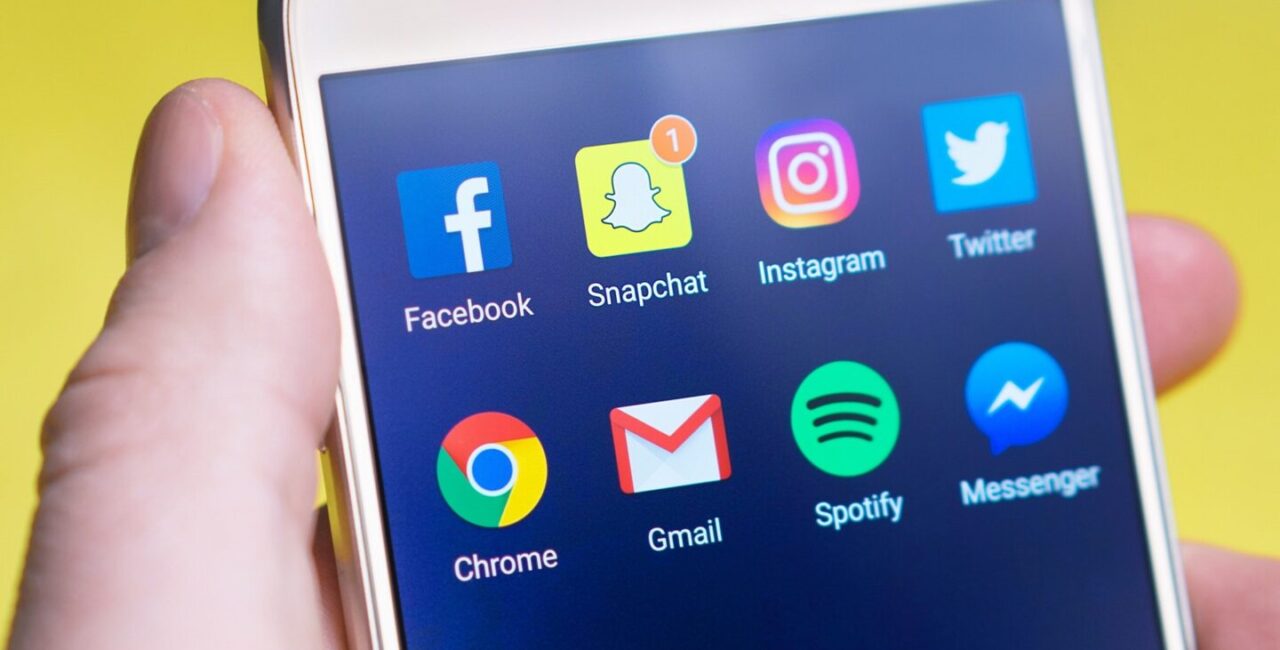 There is a screen of a white mobile phone with icons of the most popular social media Instagram, Twiter, Facebook, etc.