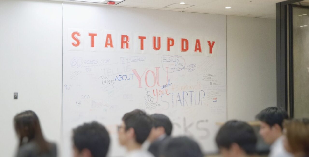 The group of people is learning in the office with a whiteboard on the wall with caption Startupday