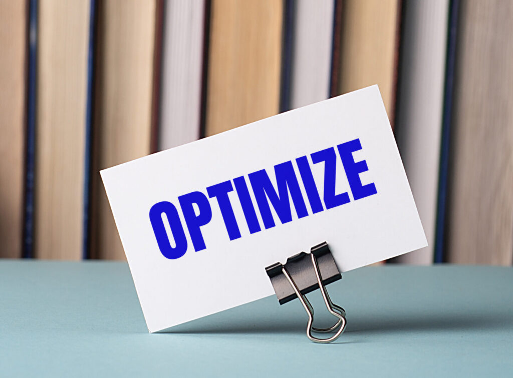 A white card with the text OPTIMIZE stands on a clip for papers on the table against the background of books.