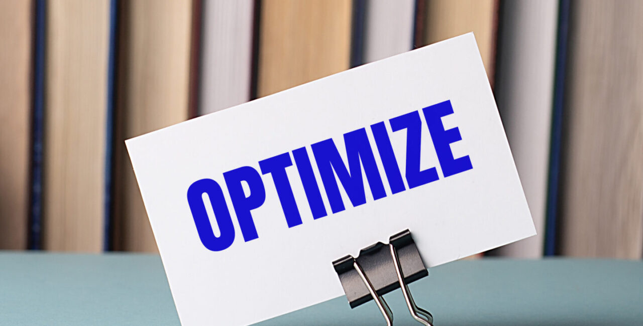 A white card with the text OPTIMIZE stands on a clip for papers on the table against the background of books.