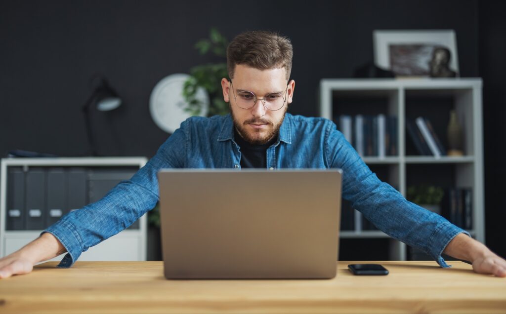 Confident young man in eyewear focused on laptop screen, business owner thoughts