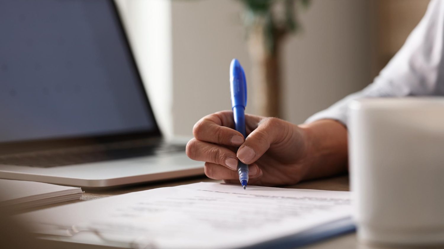 A hand holding a pen over a printed document, with an open laptop and a cup beside it.