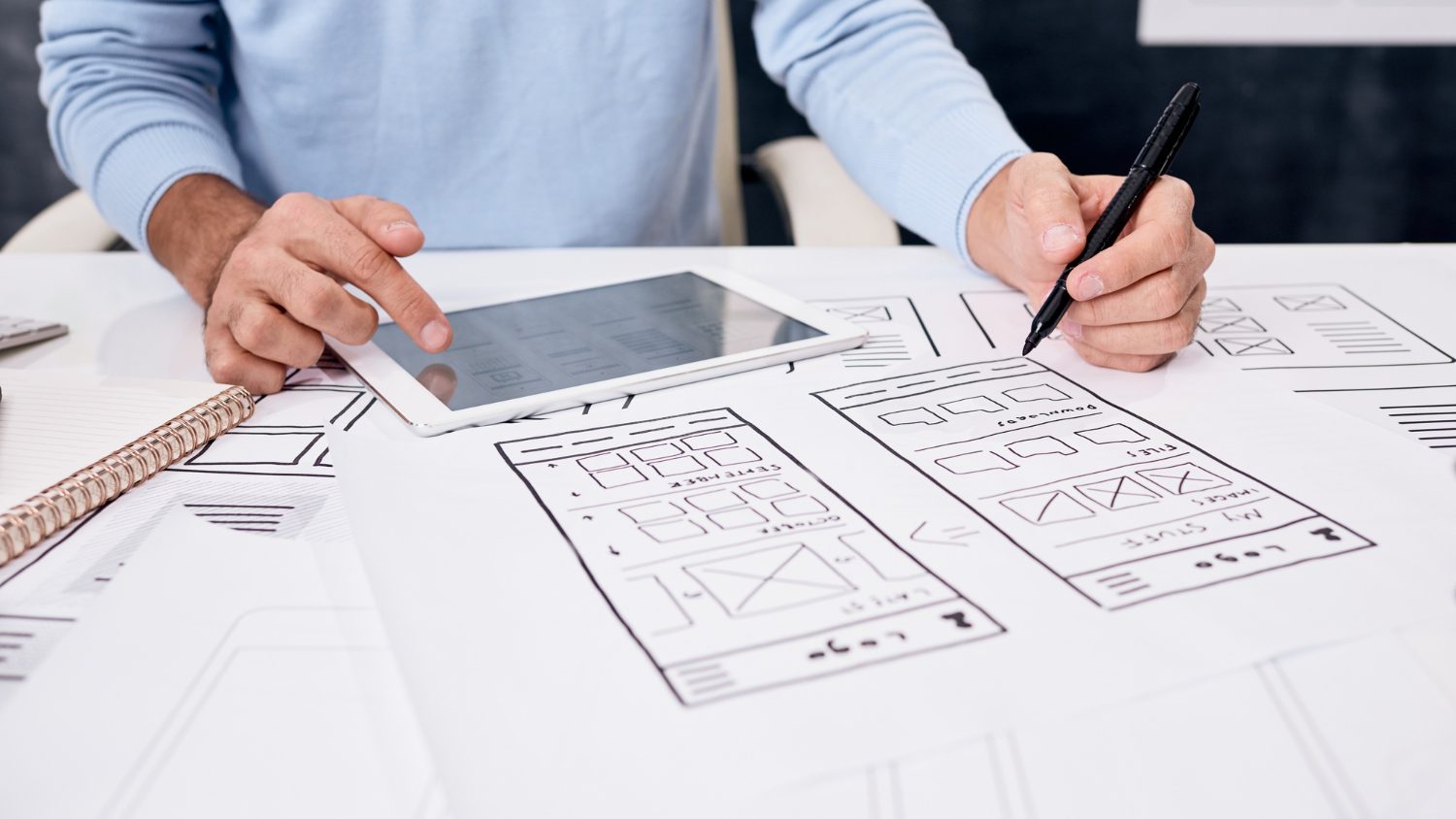 A person at a table sketching mobile page wireframes on paper, with a tablet showing a grid layout beside them.