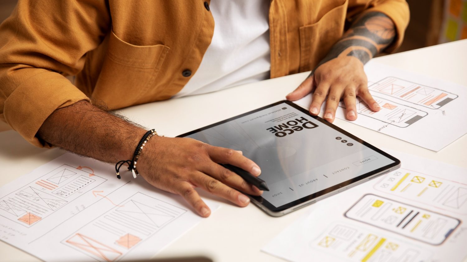 A person reviewing a home page mockup on a tablet, with paper wireframes for phone screens spread around it.